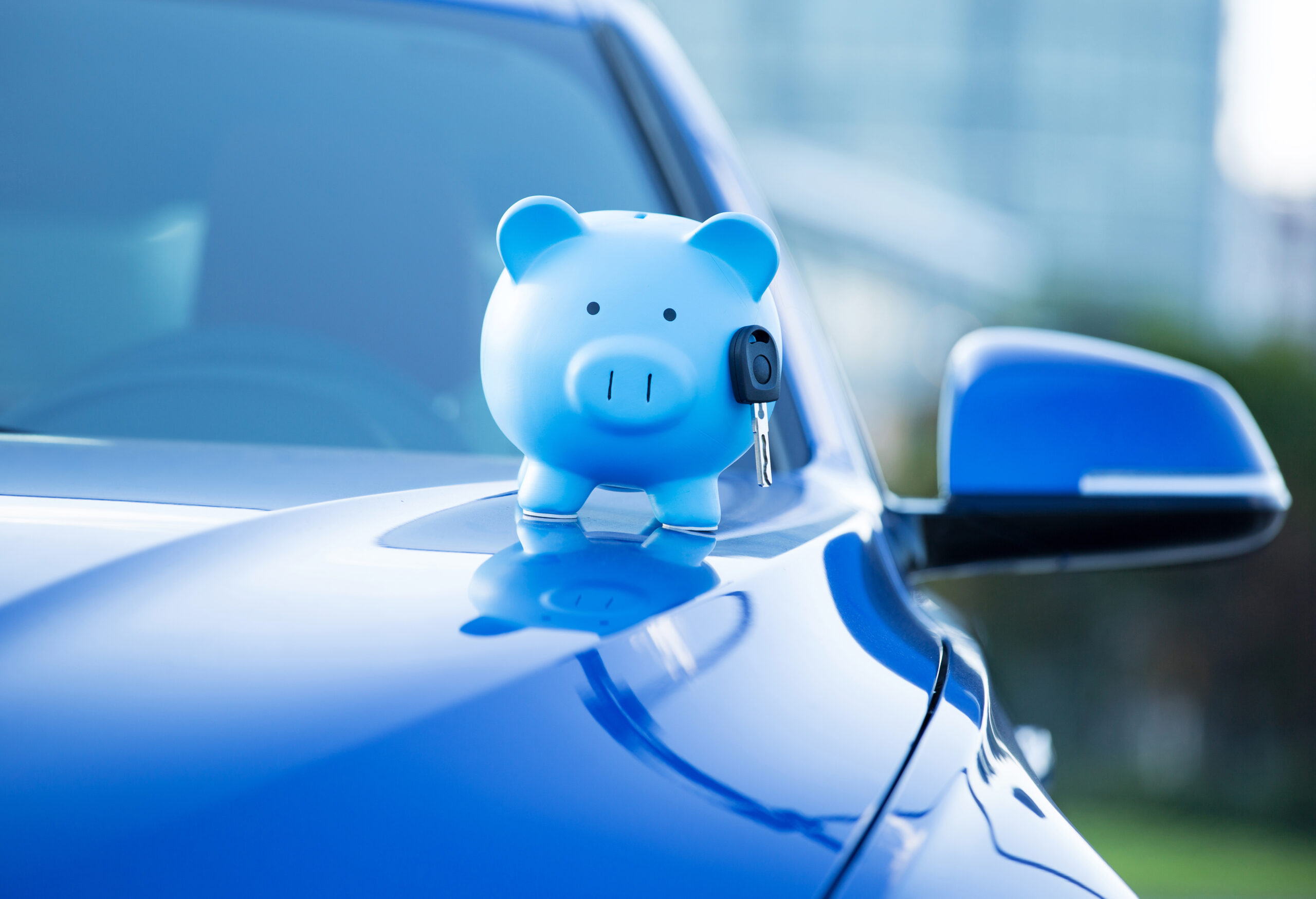 Closeup cropped image blue new car with piggy bank and key on hood, isolated outside corporate building